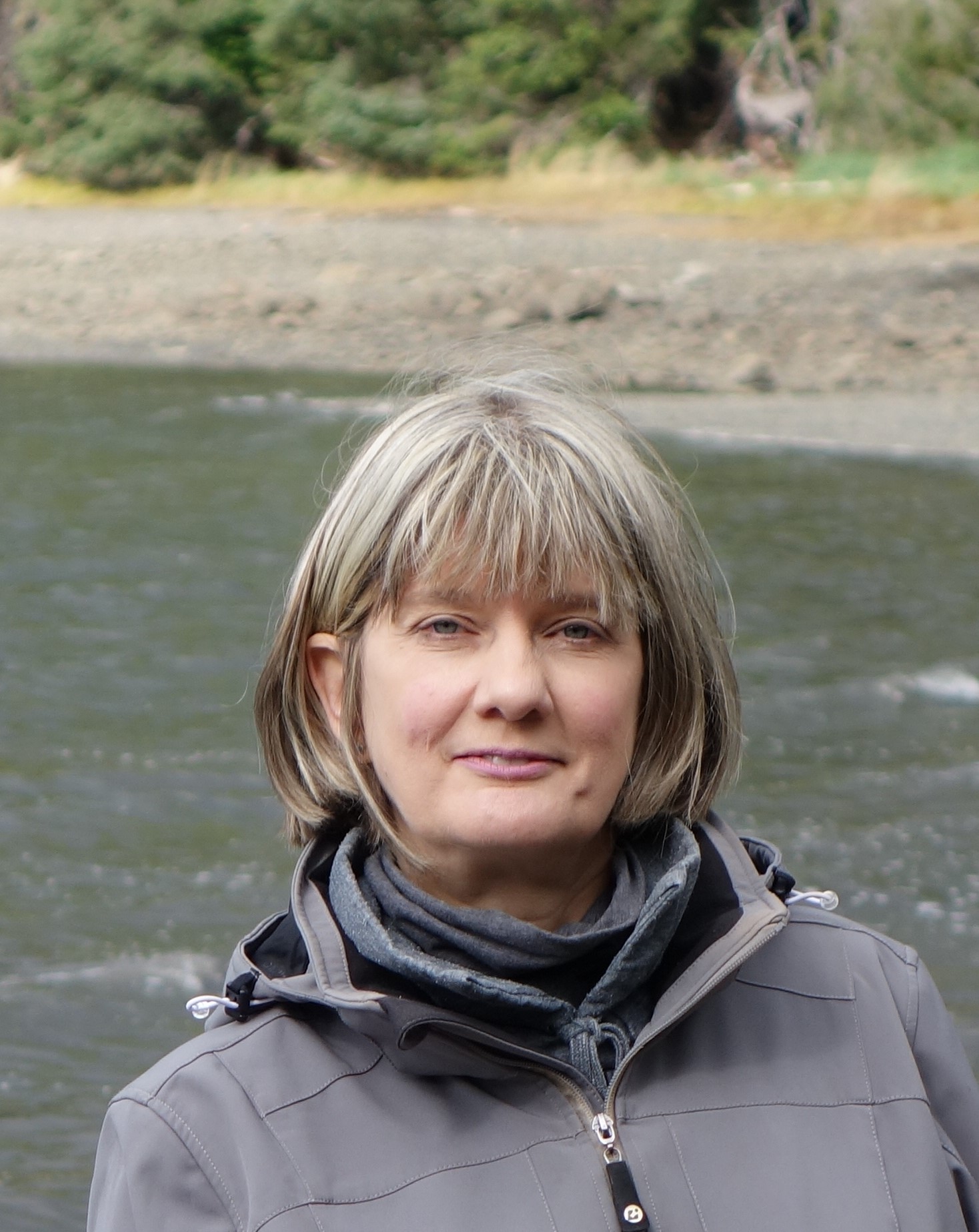 person with blond hair standing near water in a grey jacket 